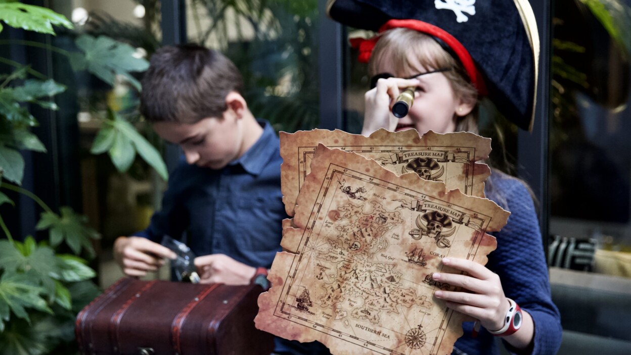Deux enfants dehors en train de jouer au pirate. La fille est déguisée en pirate et tient une carte à la main. Le garçon a un coffre sur les genoux.